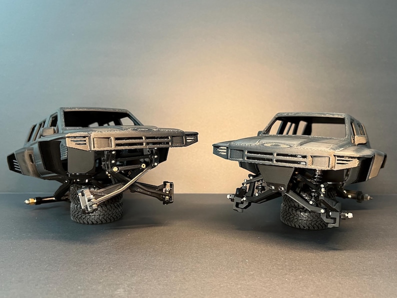 May include: Two dark gray 3D-printed model truck bodies featuring a detailed front grille, hood, and cabin structure. Each model is mounted on a single black tire, showcasing the exposed front suspension components, control arms, and steering knuckles.