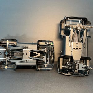 May include: Two black plastic model vehicle chassis components shown from an underside perspective. Each piece features intricate mechanical details including suspension arms, axles, and structural framing, displaying the internal assembly of a scale model.