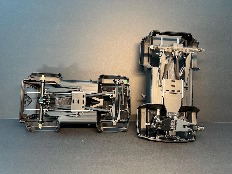 May include: Two black plastic model vehicle chassis components shown from an underside perspective. Each piece features intricate mechanical details including suspension arms, axles, and structural framing, displaying the internal assembly of a scale model.