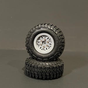 Könnte beinhalten: Zwei schwarze Crawler-Reifen mit weißen Felgen sind übereinander gestapelt. Die Reifen haben ein tiefes Profil für den Geländeeinsatz. Die Felgen haben mehrere kleine Löcher um die Mitte.