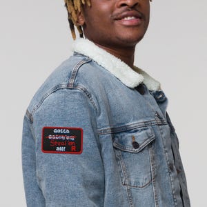 May include: A light blue denim jacket with a white sherpa collar and a rectangular patch on the sleeve. The patch is black with a red border and features the text "Gotta Catch 'em all!" with "Catch 'em" crossed out in red, and the word "Steal 'em all!" below it.