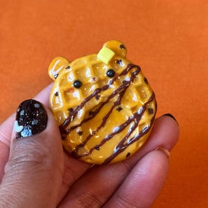 May include: A round, golden-brown waffle-shaped charm featuring a bear-like face with two small ears, black eyes, and a tiny nose. The surface is drizzled with dark brown chocolate syrup and topped with a small, square yellow butter pat.