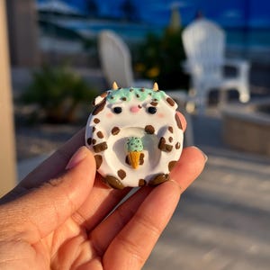 May include: A small, round, white ceramic trinket dish featuring a brown cow-spotted pattern, two small horns, and a mint green textured top. A miniature ice cream cone with mint green scoops is attached to the center of the dish.