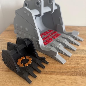 May include: Two 3D-printed excavator bucket models. One is gray with red text reading "PEKA EXCAVATION REPAIR". The other is black with an orange gear emblem. Both have multiple teeth.