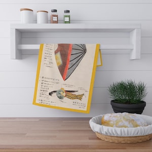 Puede incluir: Una toalla de cocina amarilla con una ilustración vintage de una mujer japonesa y un paraguas japonés tradicional. La ilustración está en tinta negra, roja y azul. La toalla está colgada en un estante blanco.