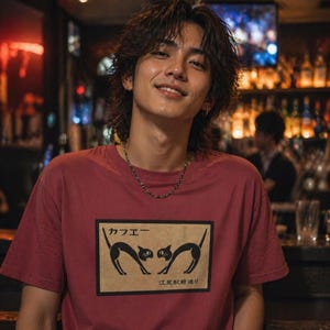 以下が含まれることがあります： 2匹の黒い猫のシルエットが向かい合っている長方形のグラフィックが特徴の、マルーン色のTシャツ。グラフィックには日本語のテキストが含まれています。画像内の人物はシルバーのチェーンネックレスを着用しています。