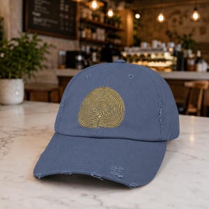 May include: A dusty blue baseball cap with a distressed, worn-in finish. The front panel features a circular, gold-coloured embroidered design consisting of a series of concentric, maze-like lines that form a stylised, symmetrical pattern.