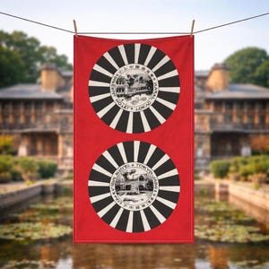 May include: Red rectangular towel with two circular black and white designs. Each design features a building illustration and text in a circular pattern. The towel is hanging on a clothesline with a blurred outdoor background.