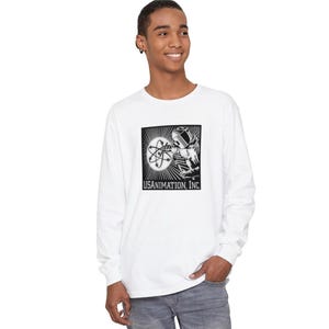 May include: White long-sleeve t-shirt with a black and white graphic of a person in a helmet holding an atom. The graphic is in a black frame with the text "USANIMATION, INC."