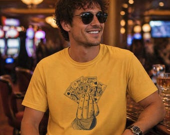 May include: Mustard yellow t-shirt with a graphic of a hand holding playing cards. The design includes spades, clubs, diamonds, and hearts. The person is wearing sunglasses and a watch.