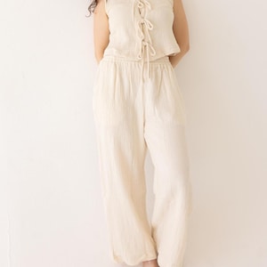 May include: A cream-colored two-piece outfit. The sleeveless top has a tie-up front. The matching pants are wide-legged. The outfit is made of a textured fabric. The model is leaning against a white wall.