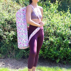 May include: A cylindrical yoga mat bag featuring a light pink fabric base with a repeating pattern of dark pink clover-like flowers and green stems. The bag includes a long, adjustable shoulder strap with a matching floral print and red trim.