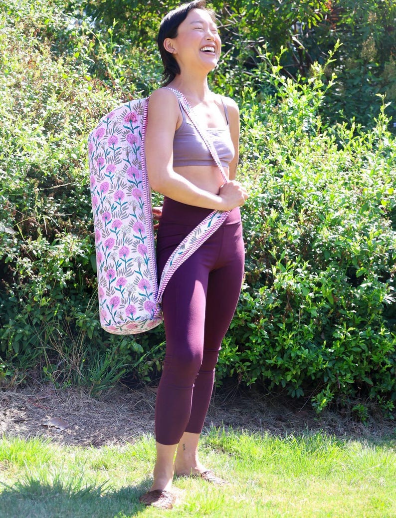 May include: A cylindrical yoga mat bag featuring a light pink fabric base with a repeating pattern of dark pink clover-like flowers and green stems. The bag includes a long, adjustable shoulder strap with a matching floral print and red trim.