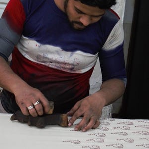 May include: A person block printing a pattern onto fabric. The person is wearing a blue, white, and red shirt. The fabric is white with a repeating pattern in a reddish-brown color. The block printing tool is made of wood.