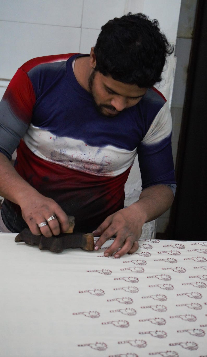 May include: A person block printing a pattern onto fabric. The person is wearing a blue, white, and red shirt. The fabric is white with a repeating pattern in a reddish-brown color. The block printing tool is made of wood.