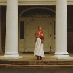 May include: A woman wearing a rust-colored jacket and a long white dress stands in front of a building with white columns. The building has a white door and an American flag is visible above.