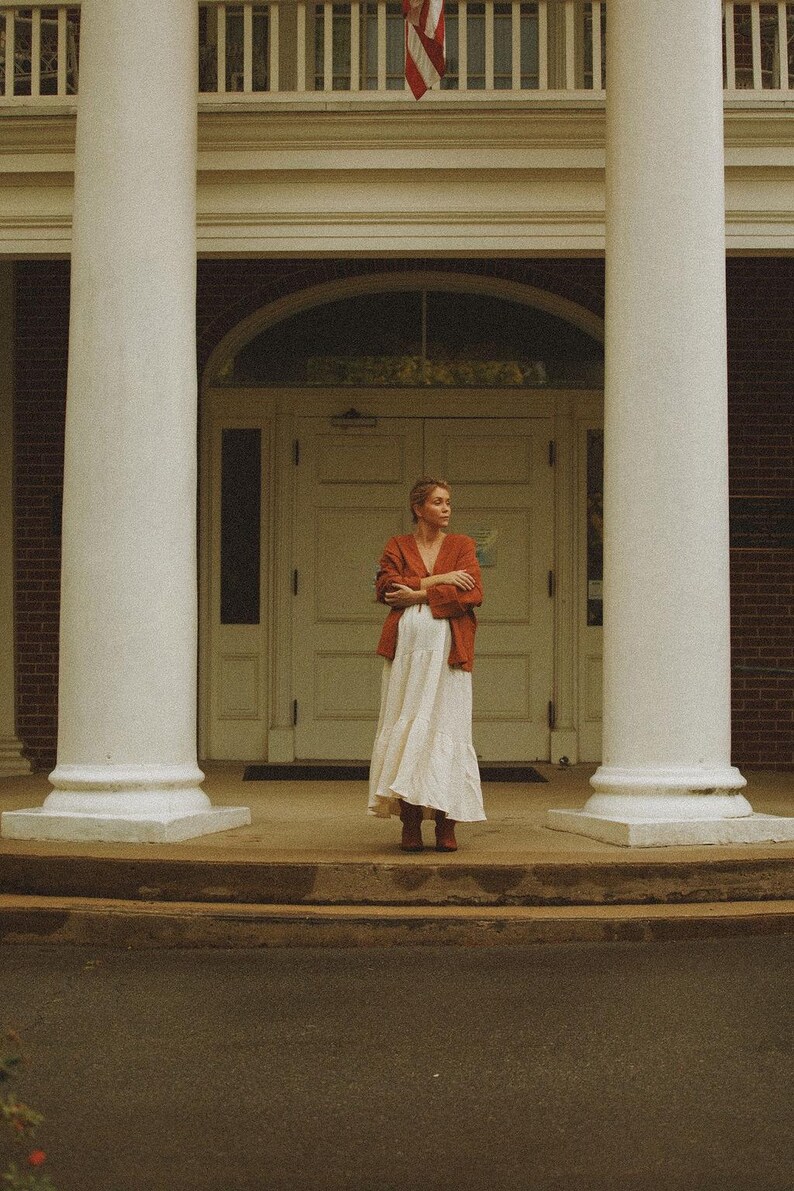 May include: A woman wearing a rust-colored jacket and a long white dress stands in front of a building with white columns. The building has a white door and an American flag is visible above.