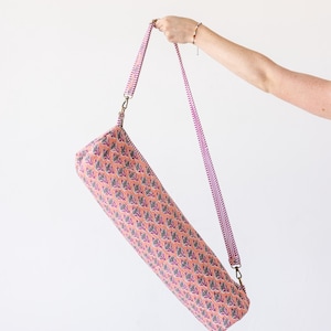 May include: A cylindrical yoga mat bag featuring a peach-colored fabric with a repeating pattern of small blue and purple floral motifs. The bag includes a long, adjustable shoulder strap with a pink and white striped design and brass hardware.