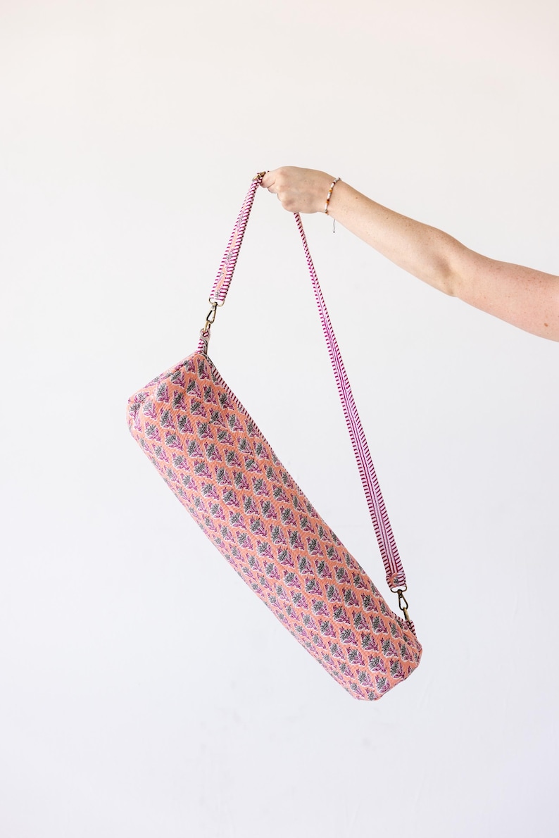 May include: A cylindrical yoga mat bag featuring a peach-colored fabric with a repeating pattern of small blue and purple floral motifs. The bag includes a long, adjustable shoulder strap with a pink and white striped design and brass hardware.