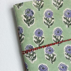 May include: A floral patterned journal with a light green background and purple flowers. The journal is bound with a red, yellow, and black string. The design is repeated across the cover.