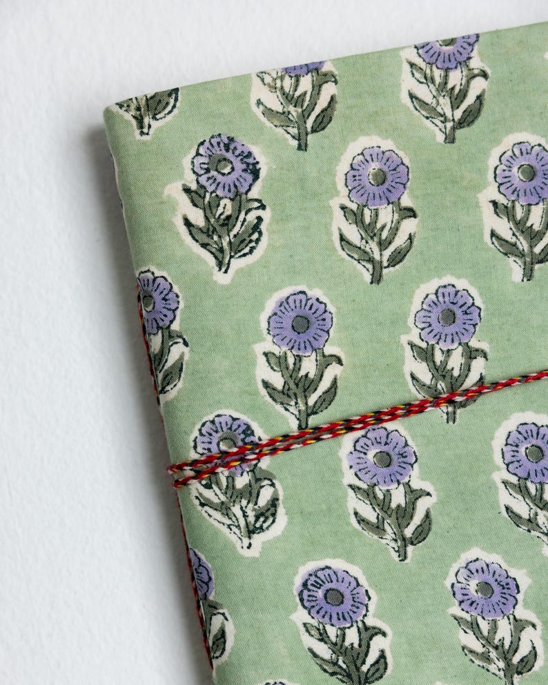 May include: A floral patterned journal with a light green background and purple flowers. The journal is bound with a red, yellow, and black string. The design is repeated across the cover.