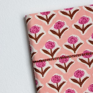 May include: A pink floral patterned notebook with a repeating design of pink flowers with brown stems and leaves. The notebook is bound with a red, black, and white string.