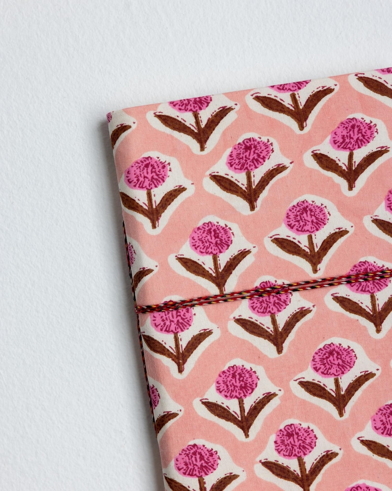 May include: A pink floral patterned notebook with a repeating design of pink flowers with brown stems and leaves. The notebook is bound with a red, black, and white string.