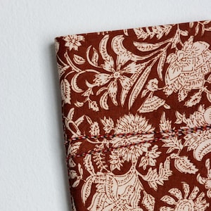 May include: Close-up of a section of fabric with a floral pattern. The fabric is a deep brown color with a cream-colored floral design. The edges are stitched with red, black, and white thread. The design features large flowers and leaves.