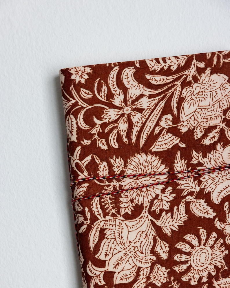 May include: Close-up of a section of fabric with a floral pattern. The fabric is a deep brown color with a cream-colored floral design. The edges are stitched with red, black, and white thread. The design features large flowers and leaves.