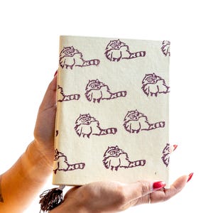 May include: A cream-colored journal featuring a repeating pattern of raccoon illustrations in a dark purple hue. The journal has a tassel attached to the spine. The design is whimsical and suitable for writing or sketching.