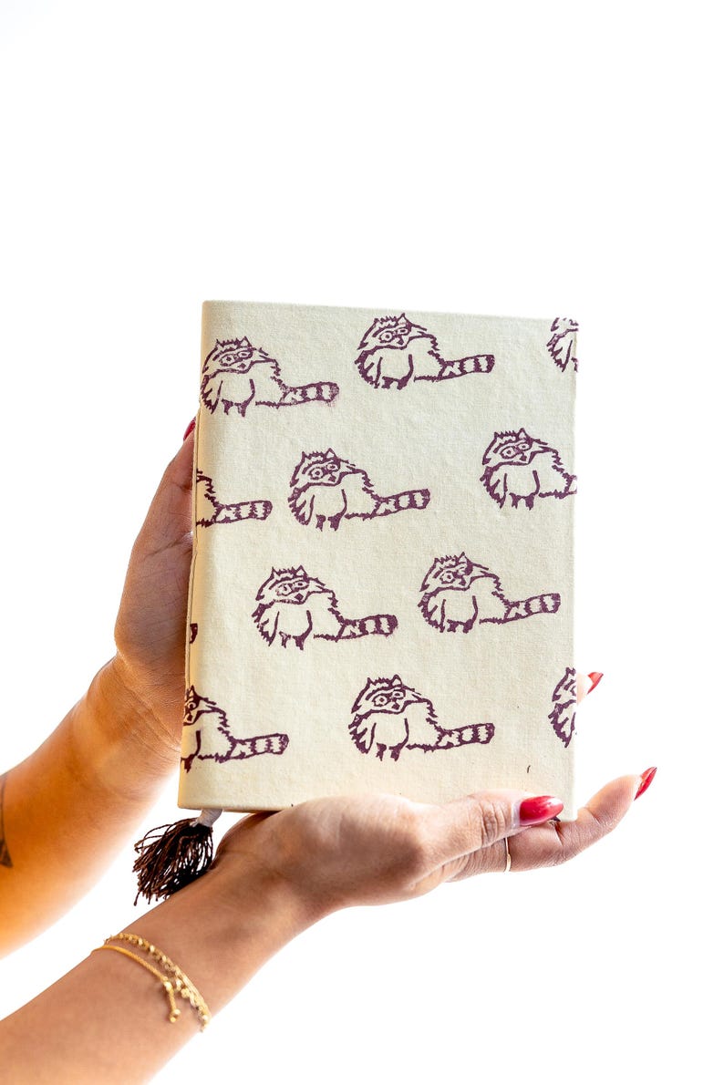 May include: A cream-colored journal featuring a repeating pattern of raccoon illustrations in a dark purple hue. The journal has a tassel attached to the spine. The design is whimsical and suitable for writing or sketching.