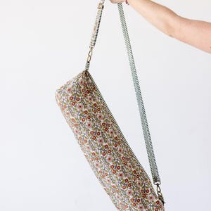 May include: A cylindrical yoga mat bag featuring a repeating floral pattern with small red flowers, green leaves, and thin brown vines on a cream background. The bag includes a long, adjustable green and white striped shoulder strap with metal clips.