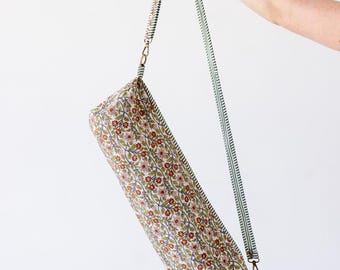 May include: A cylindrical yoga mat bag featuring a repeating floral pattern with small red flowers, green leaves, and thin brown vines on a cream background. The bag includes a long, adjustable green and white striped shoulder strap with metal clips.