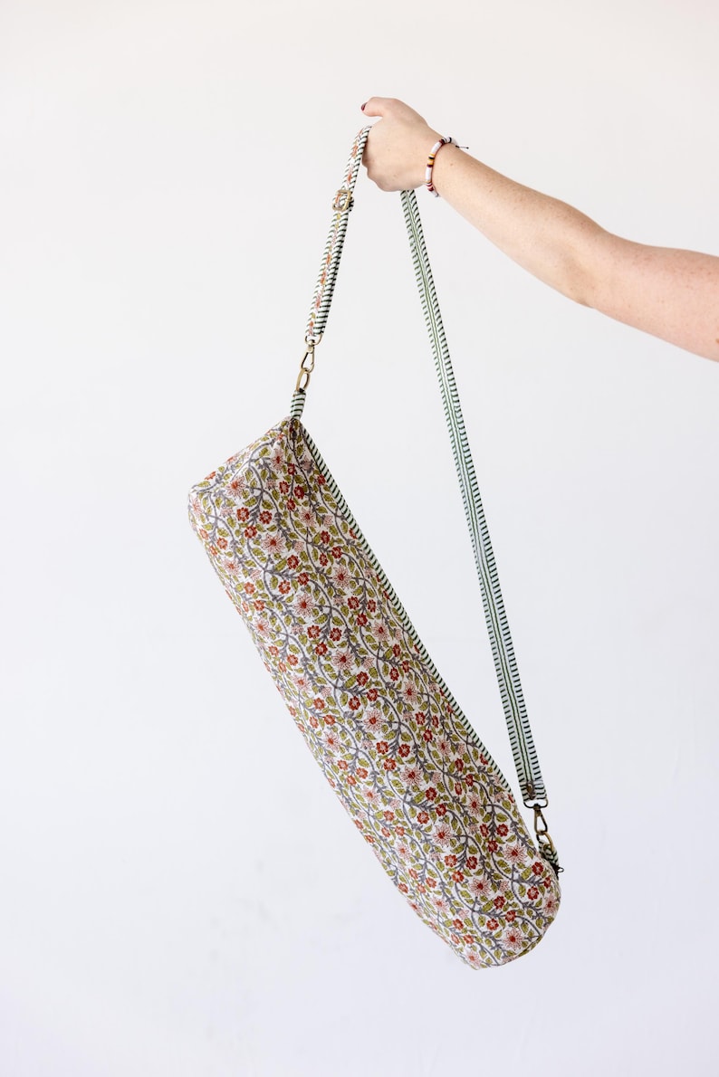 May include: A cylindrical yoga mat bag featuring a repeating floral pattern with small red flowers, green leaves, and thin brown vines on a cream background. The bag includes a long, adjustable green and white striped shoulder strap with metal clips.