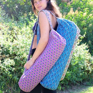 May include: A cylindrical yoga mat bag featuring a vibrant pink fabric with a repeating small floral pattern, paired with a blue and white striped shoulder strap. The bag has a zippered closure and a structured, elongated shape for carrying mats.