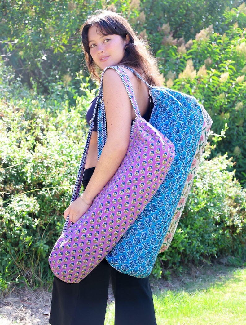 May include: A cylindrical yoga mat bag featuring a vibrant pink fabric with a repeating small floral pattern, paired with a blue and white striped shoulder strap. The bag has a zippered closure and a structured, elongated shape for carrying mats.