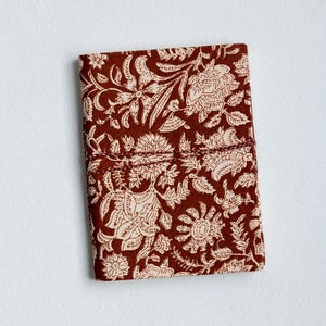 May include: A closed, rectangular journal with a floral pattern. The cover is a deep reddish-brown with a cream-colored floral design. A matching cord wraps around the journal to keep it closed. The journal is on a white background.