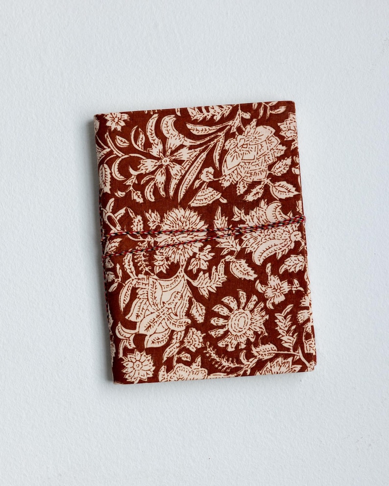 May include: A closed, rectangular journal with a floral pattern. The cover is a deep reddish-brown with a cream-colored floral design. A matching cord wraps around the journal to keep it closed. The journal is on a white background.