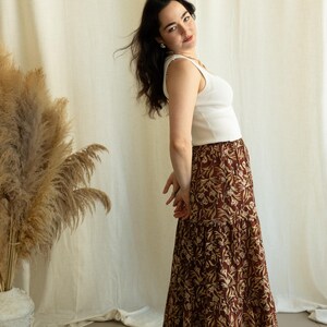 May include: A woman wearing a white ribbed tank top and a long, tiered skirt. The skirt features a brown background with a repeating floral pattern in a lighter tan color. The outfit is casual and stylish.