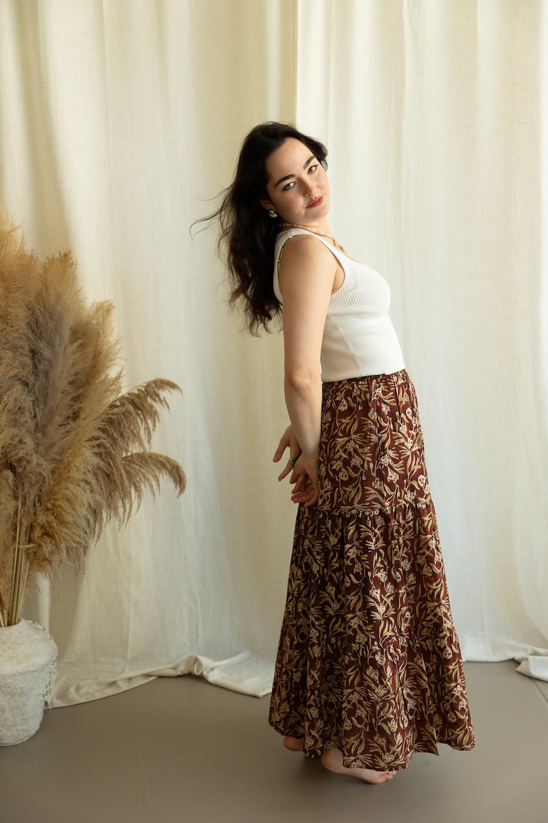 May include: A woman wearing a white ribbed tank top and a long, tiered skirt. The skirt features a brown background with a repeating floral pattern in a lighter tan color. The outfit is casual and stylish.