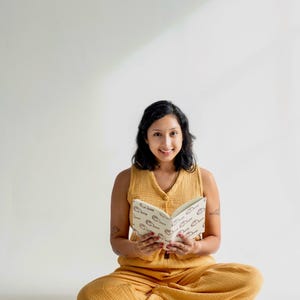 May include: A woman in a mustard yellow outfit sits cross-legged, holding an open book with a repeating pattern. Several similar books are stacked nearby. The book and outfit share a similar color palette.