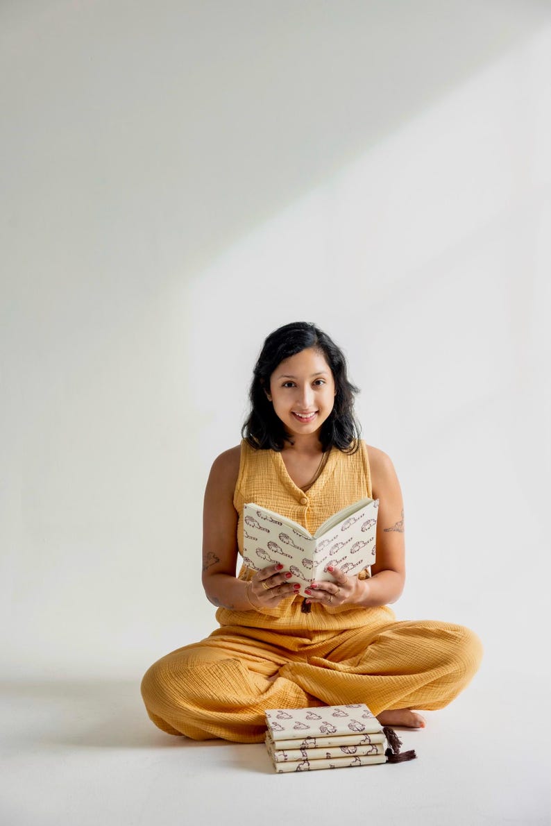 May include: A woman in a mustard yellow outfit sits cross-legged, holding an open book with a repeating pattern. Several similar books are stacked nearby. The book and outfit share a similar color palette.