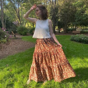 May include: A woman wearing a white sleeveless top and a tiered, floor-length skirt. The skirt is a rust color with a repeating floral pattern in a lighter shade. The woman is standing on a grassy area in front of trees.