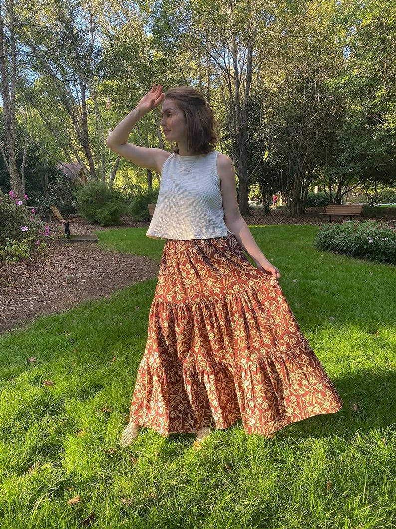 May include: A woman wearing a white sleeveless top and a tiered, floor-length skirt. The skirt is a rust color with a repeating floral pattern in a lighter shade. The woman is standing on a grassy area in front of trees.