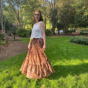 May include: A woman wearing a white sleeveless top and a long, tiered skirt. The skirt is a warm reddish-brown color with a repeating floral pattern in a lighter beige. The outfit is worn outdoors in a grassy area with trees in the background.