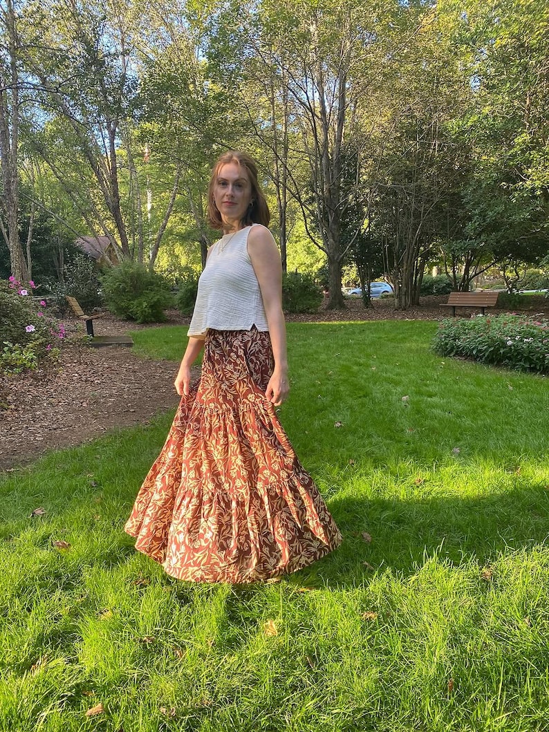 May include: A woman wearing a white sleeveless top and a long, tiered skirt. The skirt is a warm reddish-brown color with a repeating floral pattern in a lighter beige. The outfit is worn outdoors in a grassy area with trees in the background.