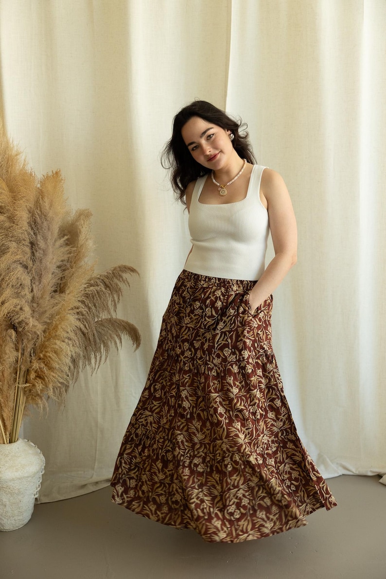 May include: A woman wearing a white ribbed tank top and a tiered, flowing skirt. The skirt features a brown background with a repeating floral pattern in a lighter beige color. The outfit is paired with a pearl necklace and earrings.