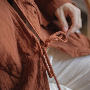 May include: Close-up of a rust-colored linen wrap top with long sleeves and a tie closure. The fabric appears soft and textured. The garment is worn over white pants.