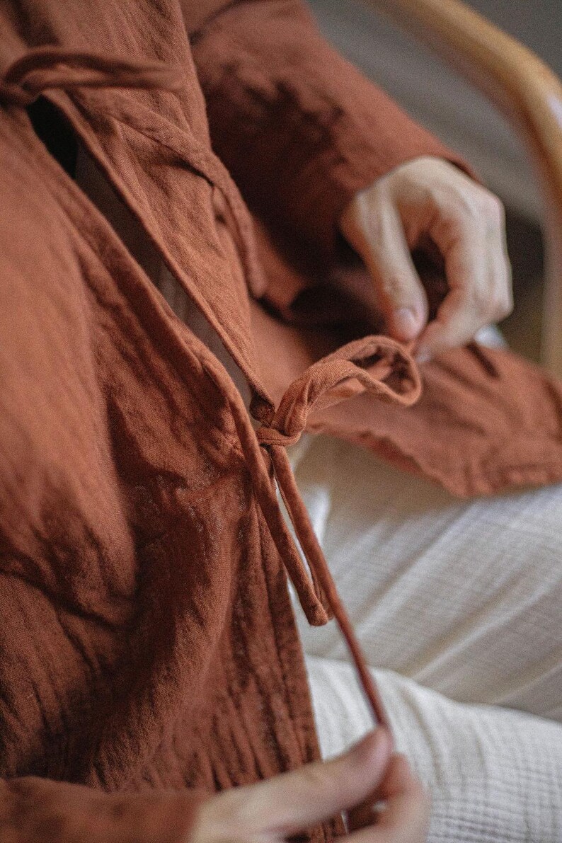 May include: Close-up of a rust-colored linen wrap top with long sleeves and a tie closure. The fabric appears soft and textured. The garment is worn over white pants.