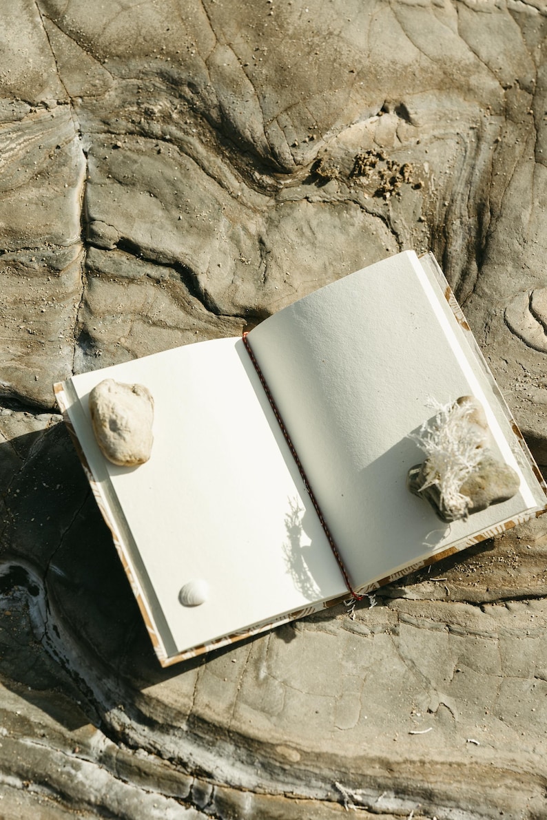 May include: An open notebook with blank white pages, resting on a textured gray rock surface. A small rock and a seashell are placed on the left page, and a rock with dried plant material is on the right. The notebook's spine is red.
